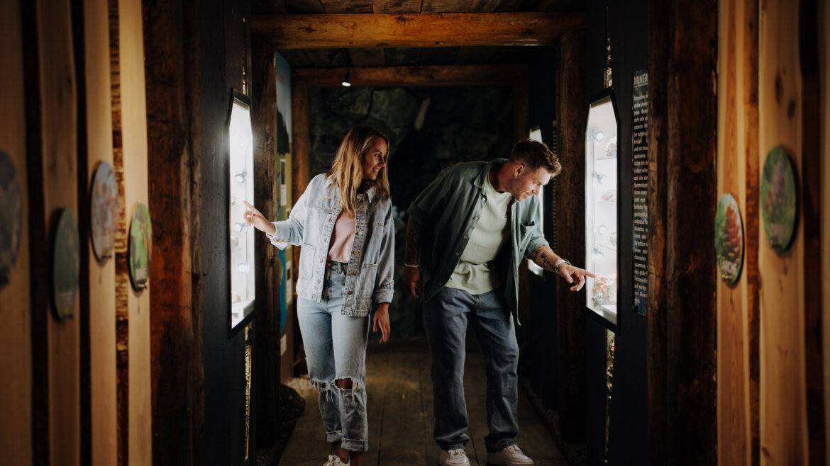 © Tourismusverband Bischofshofen | two minds Studio Zwei Personen in Bischofshofen lächeln und interagieren mit beleuchteten Displays in einem Holzgang. (vergrößerte Ansicht)