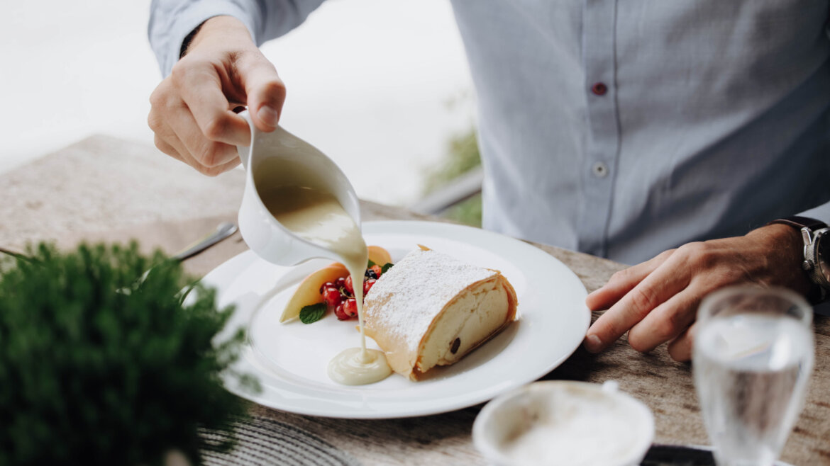 Eine Person gießt Vanillesoße über einen Topfenstrudel an einem Tisch. (vergrößerte Ansicht)