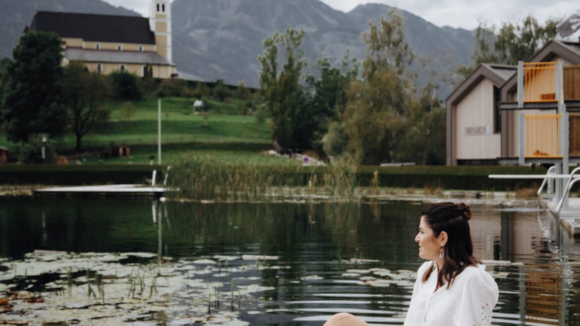 Eine Frau in Weiß sitzt auf einem Steg an einem ruhigen See, im Hintergrund die Kirche von St. Veit und die Berge. (vergrößerte Ansicht)