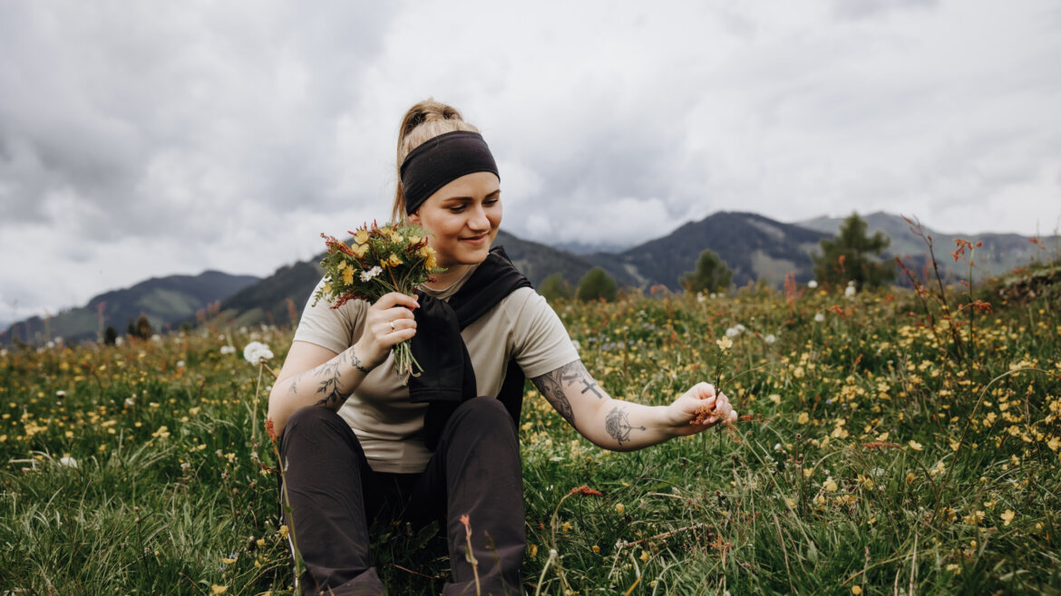 Frau, die auf einem Feld in St. Veit sitzt, einen Blumenstrauß hält und lächelt, mit Bergen im Hintergrund. (vergrößerte Ansicht)