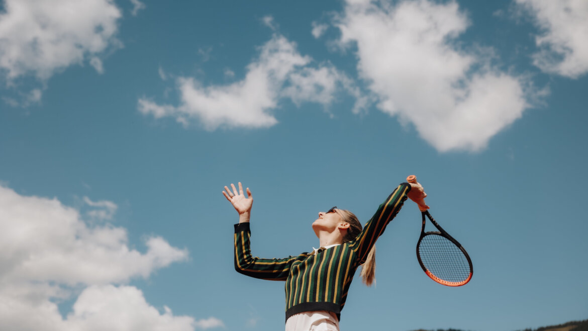 Frau in gestreiftem Oberteil serviert einen Tennisball im Freien auf einer Sonnenterrasse mit Bergen und blauem Himmel im Hintergrund. (vergrößerte Ansicht)