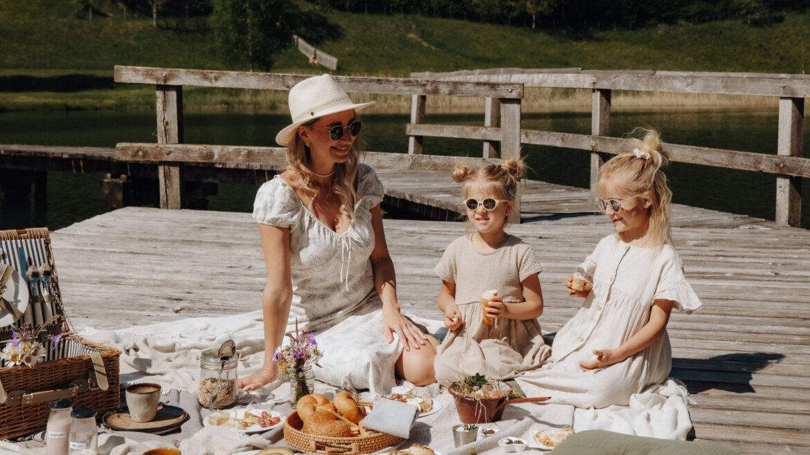Eine Frau und zwei Mädchen genießen ein Picknick auf einer Sonnenterrasse am See. (vergrößerte Ansicht)