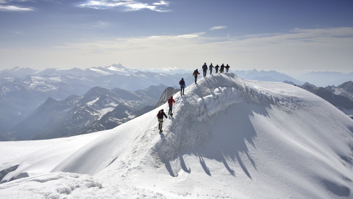 Eine Gruppe von Bergsteigern wandert in einer Reihe entlang eines verschneiten Neukirchener Bergrückens unter einem klaren blauen Himmel. (vergrößerte Ansicht)