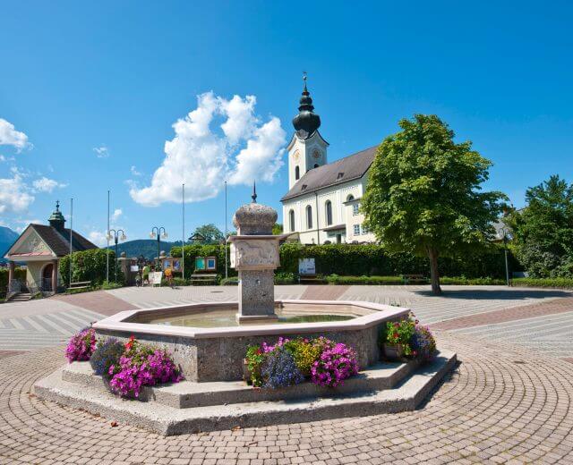 Ein Steinbrunnen mit bunten Blumen steht vor einer österreichischen Kirche in Eugendorf unter blauem Himmel.