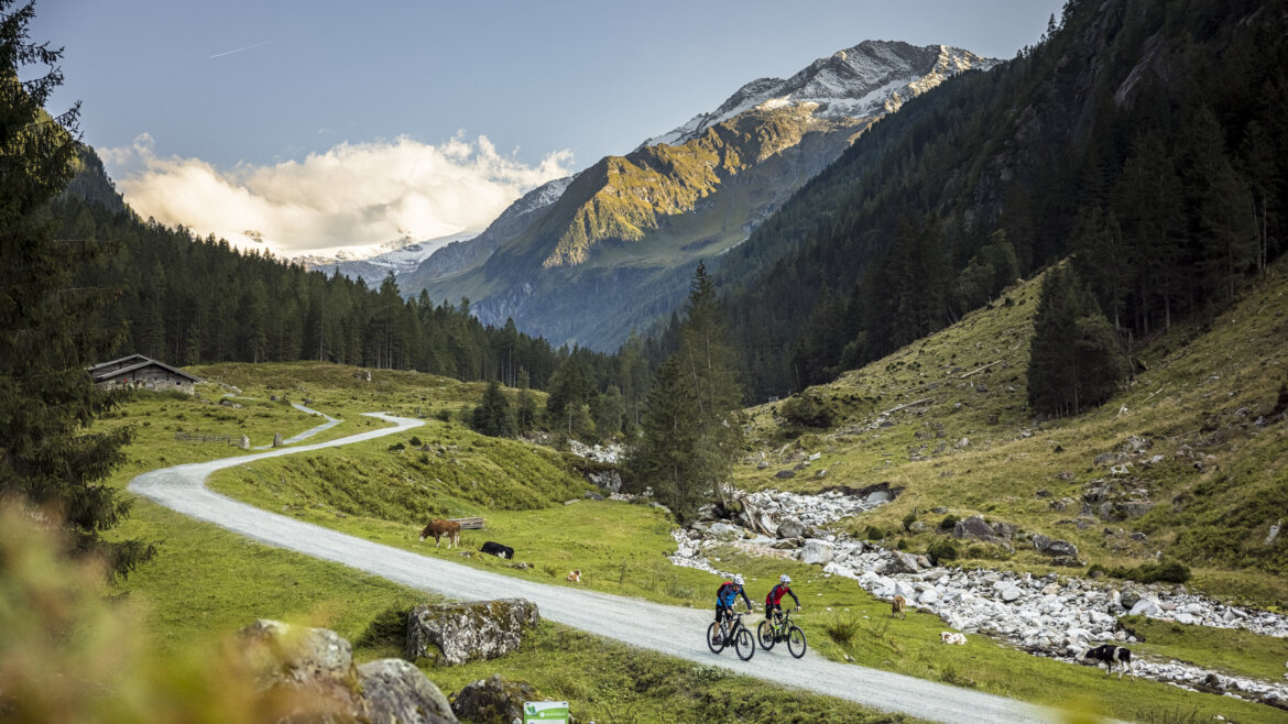 Zwei Personen fahren mit ihren Fahrrädern auf einem Radweg in Bramberg, tragen Helme und sind von neben einem Gebirgsbach und Berge im Hintergrund (vergrößerte Ansicht)