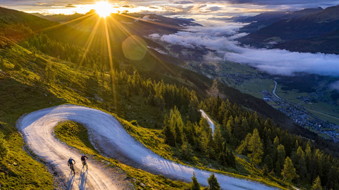 Zwei Radfahrer fahren bei Sonnenaufgang in der Nähe von Neukirchen bergauf, umgeben von Bäumen und malerischen Bergtälern. (vergrößerte Ansicht)