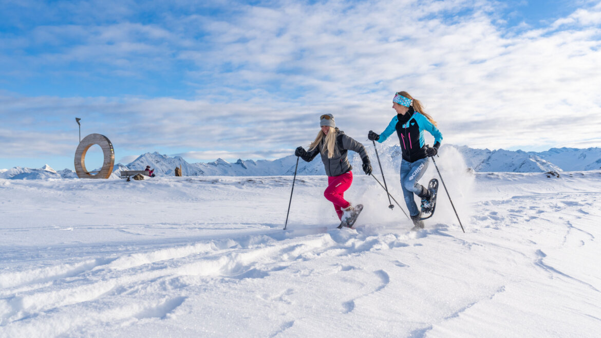Zwei Menschen, die mit ihren Schneeschuhen durch die verschneite Landschaft Neukirchens laufen, mit Bergen und blauem Himmel im Hintergrund. (vergrößerte Ansicht)