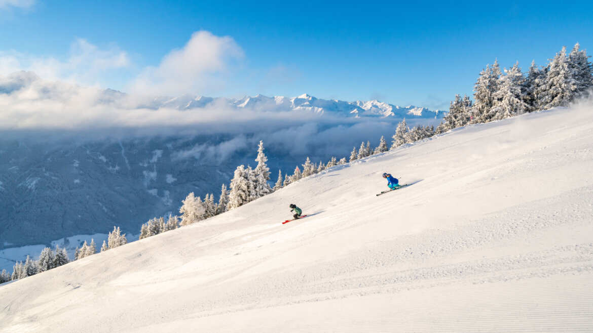 Zwei Skifahrer gleiten einen verschneiten Neukirchener Hang hinunter, umrahmt von schneebedeckten Bäumen und fernen Gipfeln unter einem blauen Himmel. (vergrößerte Ansicht)