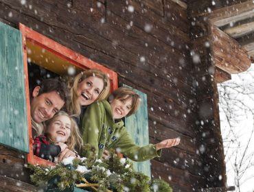 Eine lächelnde vierköpfige Familie lehnt sich aus einem verschneiten Hüttenfenster und genießt den Urlaub am Bauernhof im fallenden Schnee.