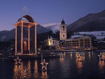 Ein Dorf am See in der Abenddämmerung in Sankt Wolfgang am See, mit sternförmigen Lichtern und einer Laterne, die sich auf dem Wasser spiegelt.
