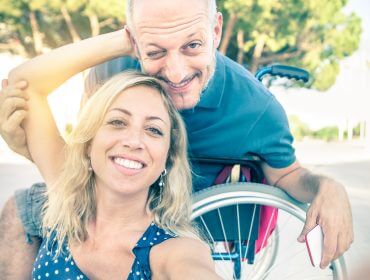 Eine lächelnde Frau macht ein Selfie mit einem Mann im Rollstuhl, der seinen barrierefreien Urlaub im Freien genießt.