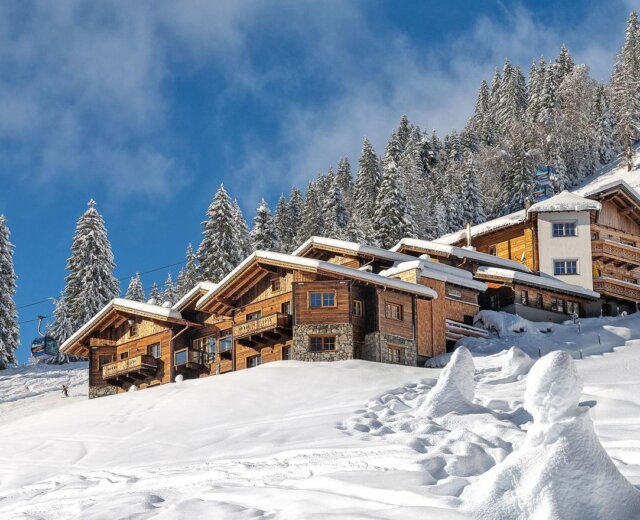 Herzwies Winter (c) Herzwies Almdörfer-Holzhütten in verschneiten Bergen, umgeben von Kiefern und einem strahlend blauen Himmel.
