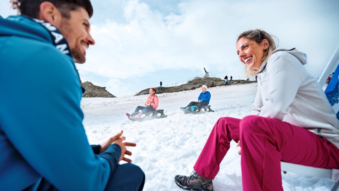 © Kitzsteinhorn - ICE ARENA Zwei Erwachsene sitzen lächelnd im Schnee, während im Hintergrund zwei Personen auf einem verschneiten Hügel Schlitten fahren. (vergrößerte Ansicht)