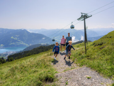 Mr-Offenblende.at_Schmitten_Social_Family_WEB-05168_Bearbeitet Eine Familie mit zwei Kindern wandert auf einem grasbewachsenen Weg auf der Schmittenhöhe und genießt den Blick auf einen See und die Berge in der Nähe einer Seilbahn.