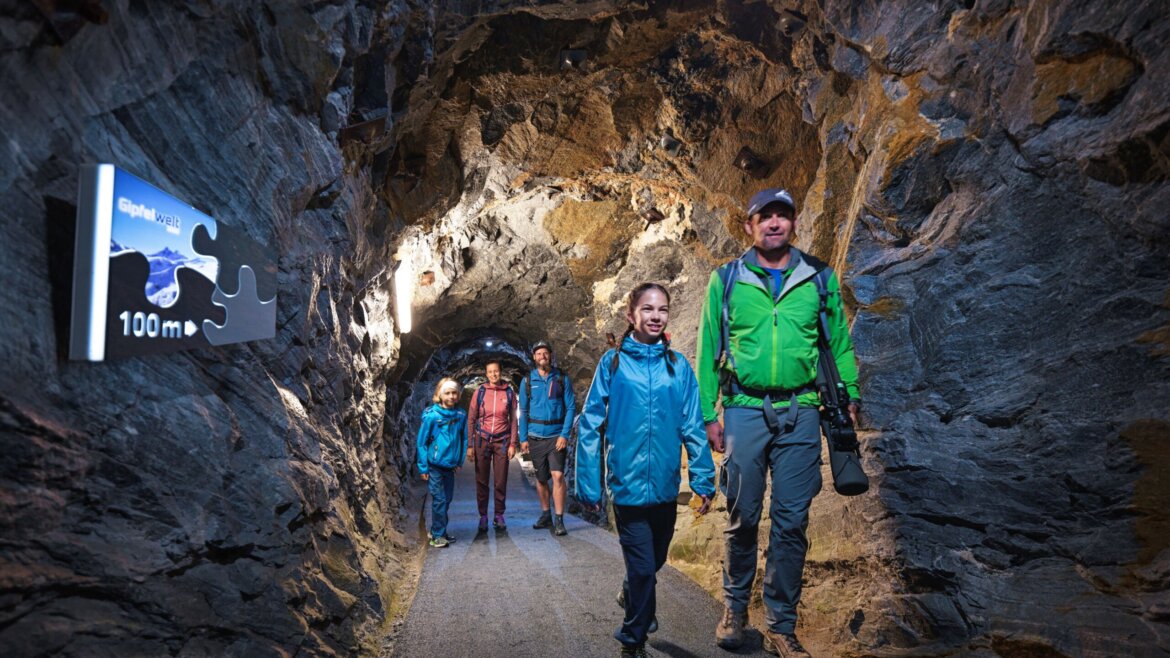 ©Kitzsteinhorn - Nationalpark Gallery Eine Familie in Wanderkleidung geht durch einen beleuchteten Tunnel mit einem Schild mit der Aufschrift "100 m" und einem Puzzleteil an der Wand. (vergrößerte Ansicht)