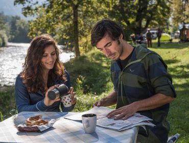 Eine Frau schenkt Kaffee ein, während ein Mann an einem Fluss liest und die Natur in der Nähe von malerischen Campingplätzen genießt.