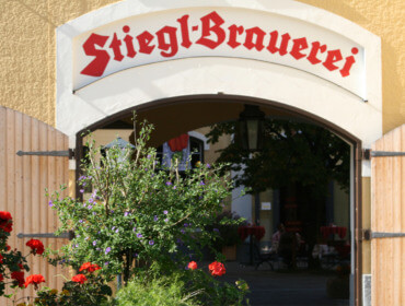 Torbogen_StieglBrauwelt Gewölbter Eingang mit Stiegl-Schild darüber und bunten Blumen im Vordergrund.