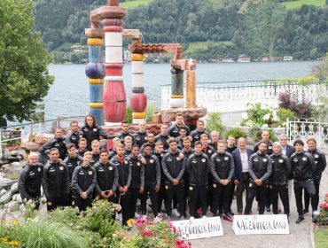 Eine Fußballmannschaft im Fußball-Trainingscamp posiert in Trainingsanzügen an einem See, einer lebendigen Skulptur und Bergen.