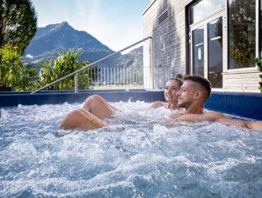 Aqua Salza (c) Aqua Salza Ein lächelndes Paar entspannt sich in einem Aqua Salza-Whirlpool im Freien mit Bergen und Grünflächen im Hintergrund.
