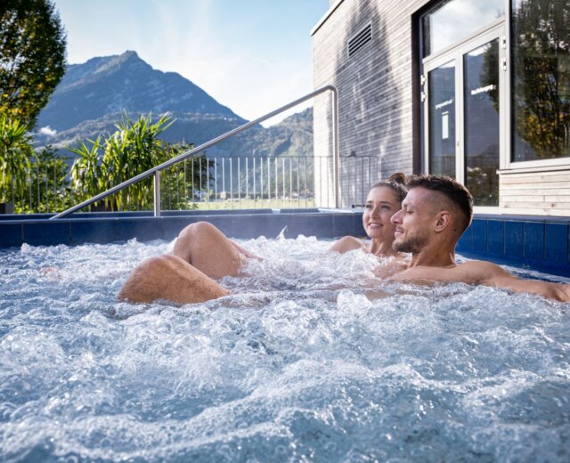Ein lächelndes Paar entspannt sich in einem Aqua Salza-Whirlpool im Freien mit Bergen und Grünflächen im Hintergrund.