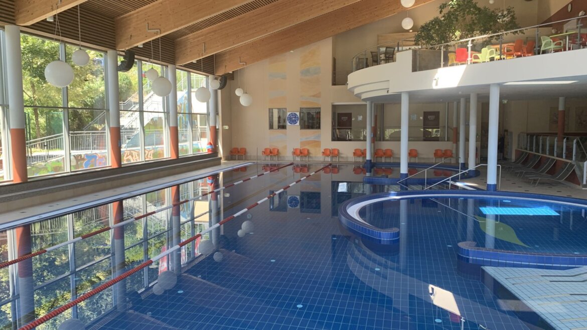 (c) Aqua Salza Hallenbad mit blauen Fliesen im Aqua Salza, große Fenster, Holzdecke, Stühle und Pflanzen im Hintergrund. (vergrößerte Ansicht)