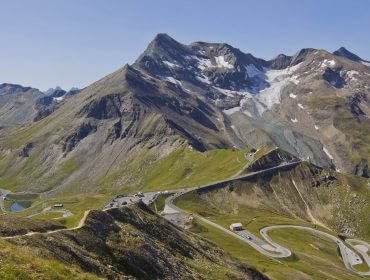 Kurvenreiche Bergstraße mit Autos, Verkehrslage klar, umgeben von grünen Hügeln und schneebedeckten Gipfeln unter einem klaren Himmel.