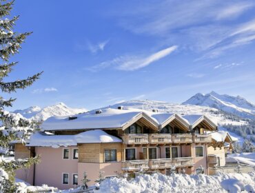 Ein schneebedecktes Gebäude im Chalet-Stil mit Balkonen vor dem Hintergrund verschneiter Berge und eines strahlend blauen Himmels.