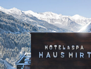 Ein modernes Hotel- und Spa-Gebäude mit dem Schriftzug HOTEL & SPA HAUSHIRT steht vor einer Kulisse aus schneebedeckten Bergen und Kiefernwäldern unter einem klaren blauen Himmel.