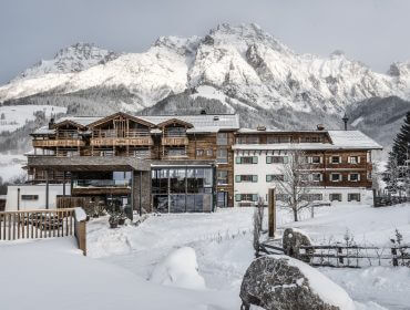 Ein großes Chalet aus Holz und Glas liegt in einer verschneiten Landschaft mit Bergen im Hintergrund.