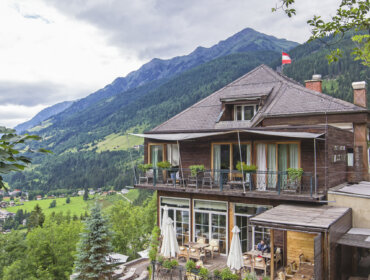 Mehrstöckiges Holzhaus mit Balkonen mit Blick auf ein üppig grünes Tal und ferne Berge unter einem bewölkten Himmel.