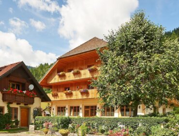 Blasiwirt Zwei charmante Landgut Blasiwirt-Holzhäuser mit Balkonen und Gärten schmiegen sich inmitten grüner Hügel unter blauem Himmel.