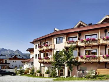 Charmantes Alpenhotel mit blumengeschmückten Balkonen, Bergblick und Menschen, die neben geparkten Autos spazieren gehen.