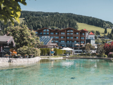 Ein großes Holzhotel mit roten Dächern liegt an einem klaren türkisfarbenen Teich, umgeben von Bäumen, Grün und Hügeln unter blauem Himmel. In der Nähe des Wassers sind Liegestühle und Sonnenschirme aufgestellt.