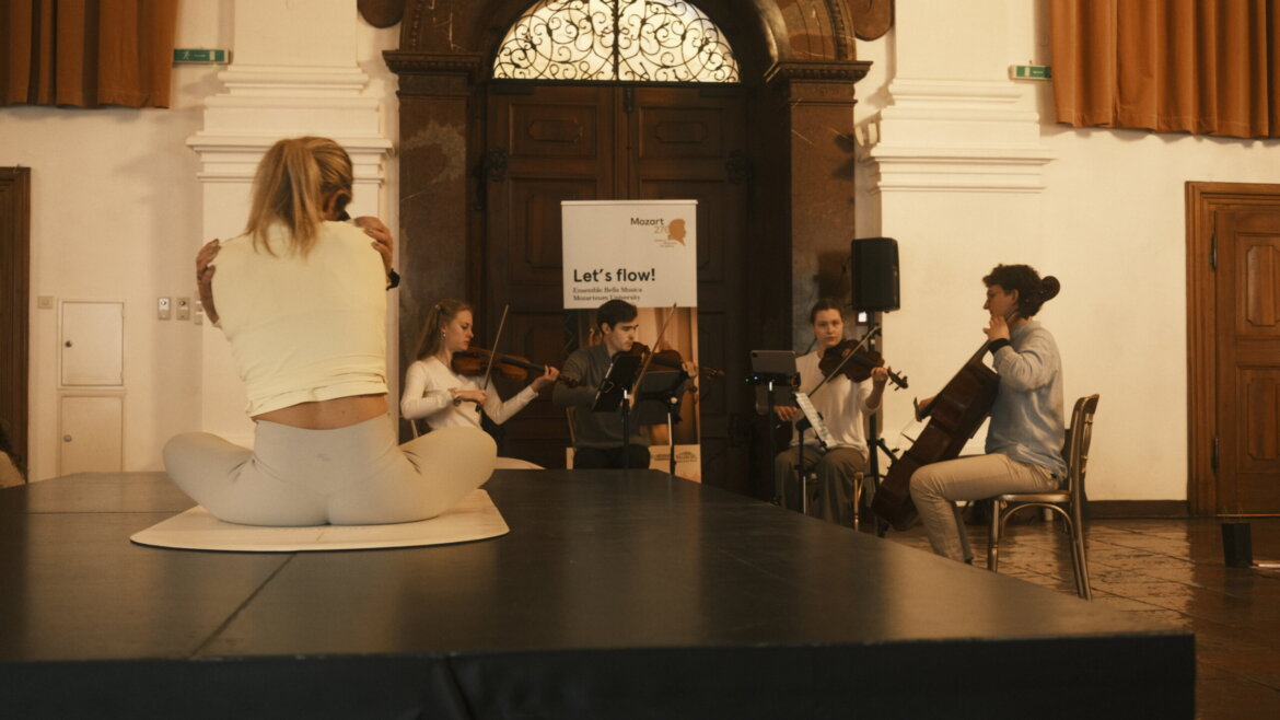 Eine Frau sitzt im Schneidersitz auf der Bühne, gegenüber von Musikern, die in einem großen Saal Streicher spielen. (vergrößerte Ansicht)