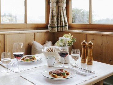 Ein gemütlicher Restauranttisch für zwei Personen mit Wein, Fleischgerichten, Blumen und Brot am Fenster.
