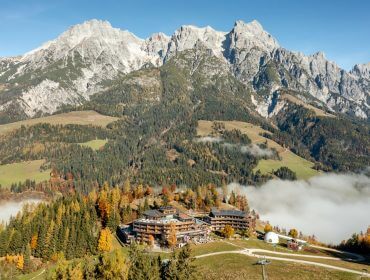 Eine von herbstlichen Bäumen umgebene Berglandschaft mit Nebel und hohen Felsgipfeln im Hintergrund.