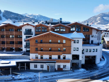 Ein großes Alpenhotel mit Holzbalkonen, umgeben von Schnee und Bergen unter blauem Himmel.