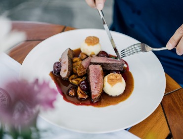 Eine Person schneidet Fleisch mit Soße, Knödeln und Gemüse auf einem weißen Teller an einem Holztisch.