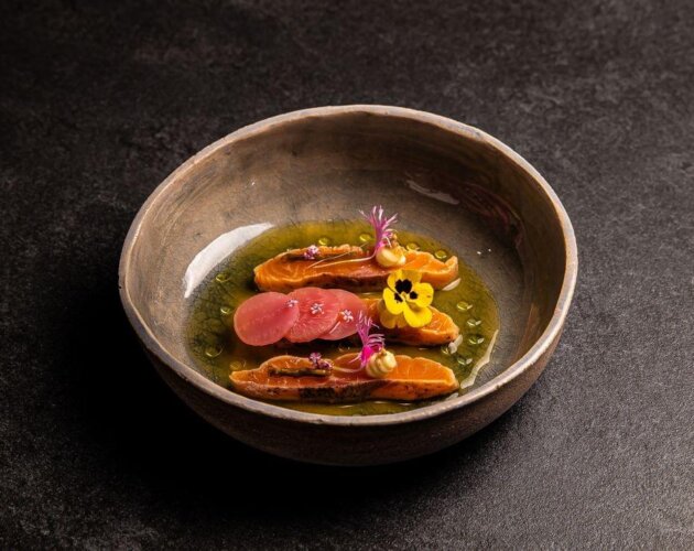Eine Keramikschale mit Haubenküche-Gourmet-Lachs, rosa Radieschenscheiben, essbaren Blüten und grüner Soße.