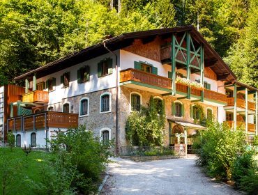 Ein rustikales, mehrstöckiges Haus mit grünen Fensterläden, Holzbalkonen und üppigem Laub drum herum.