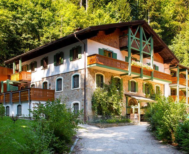 Ein rustikales, mehrstöckiges Haus mit grünen Fensterläden, Holzbalkonen und üppigem Laub drum herum.