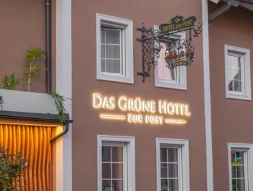 Das-grüne-Hotel-zur-Post Außenansicht des Grünen Hotels zur Post mit einem kunstvoll gestalteten Eisenschild und beleuchteten Buchstaben.