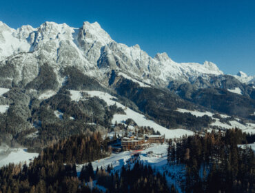 Schneebedeckte Berge und Wälder umgeben eine Hütte unter einem klaren blauen Himmel in einer Winterlandschaft.