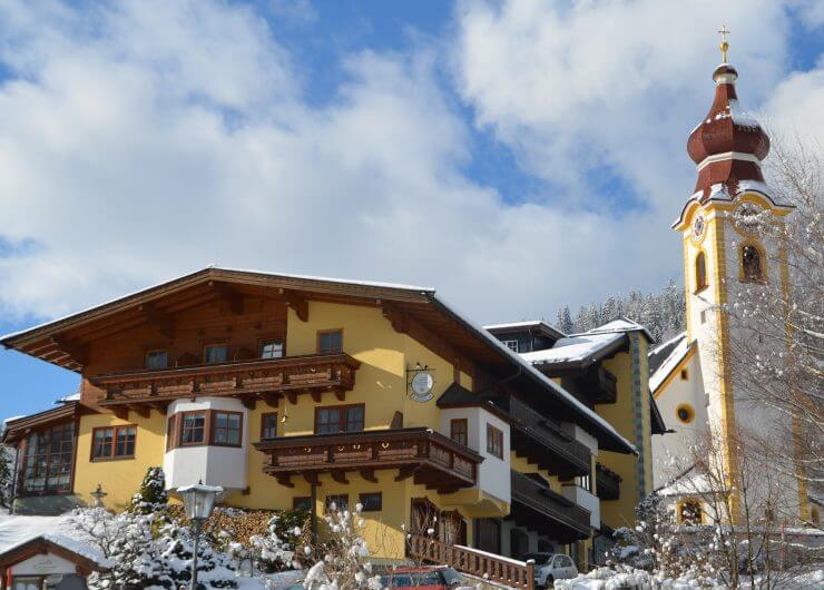 Kirchenwirt im Winter - gelbe Hausfassade, Holzbalkone, rechts dahinter die Kirche