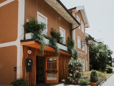 Front das Grüne Hotel zur Post Ein kleines, braunes Gebäude mit Pflanzen auf dem Balkon und einer offenen Holztür an einem sonnigen Tag.