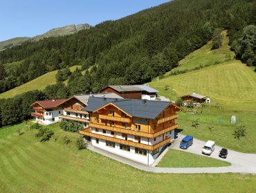 Große Holzhütte, umgeben von grünen Hügeln, Bäumen und geparkten Autos, mit Bergen im Hintergrund.