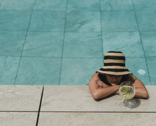 Eine Person mit gestreiftem Hut entspannt sich mit einem Getränk in der Hand am Pool des Bioparadieses im Hotel SalzburgerLand.