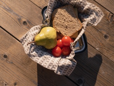 Eine Brotdose mit Brot, Kirschtomaten und einer Birne auf einem gemusterten Tuch, das auf einem Holztisch steht.