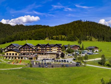Ein Panoramablick auf eine große Hütte, umgeben von grünen Hügeln, Bäumen und kurvenreichen Straßen unter blauem Himmel.