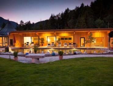 Modernes Haus mit großen Fenstern und Außenpool, wie BioVitalHotel Sommerau, warm beleuchtet in der Abenddämmerung zwischen Gärten.